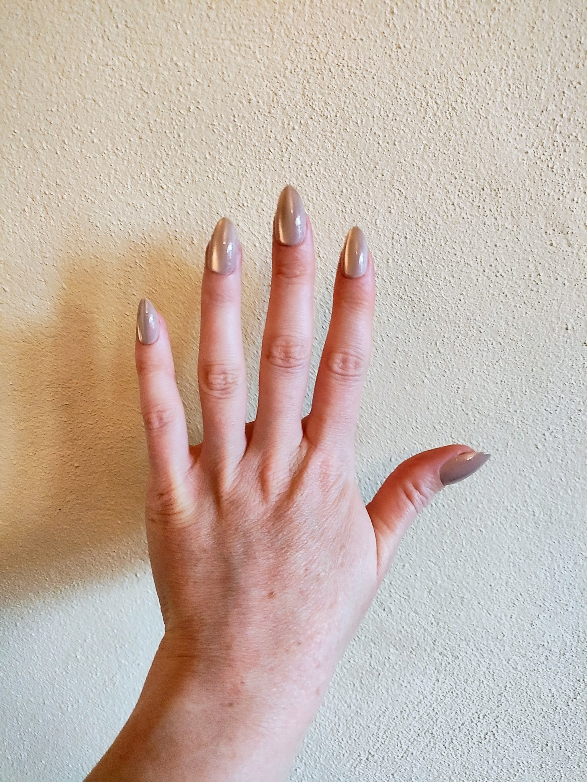 Left hand showing latte brown in our almond shape, with a slight shimmer, on a cream wall background.