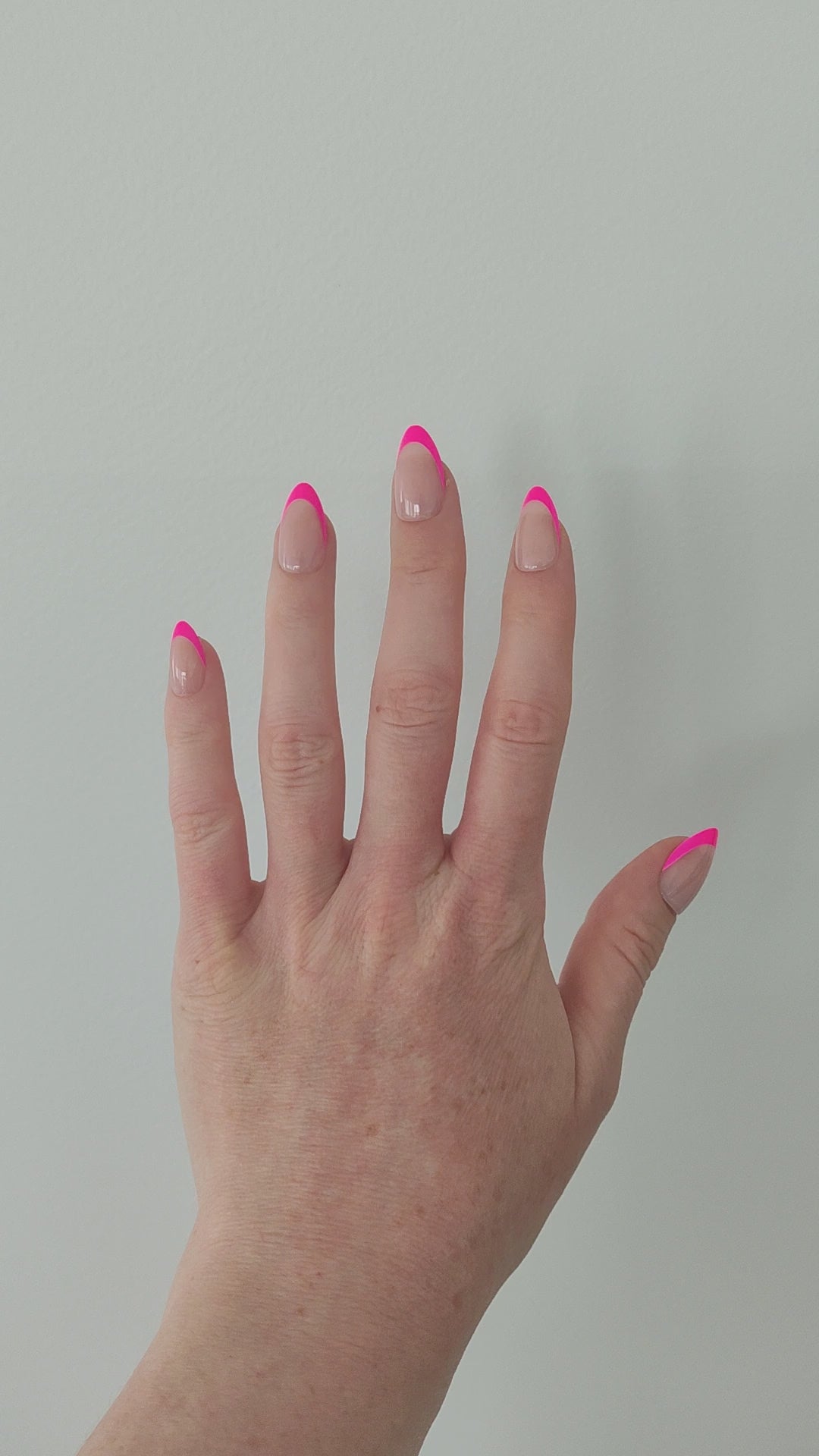 Left hand showing almond nails with a hot pink French tip on a white wall background